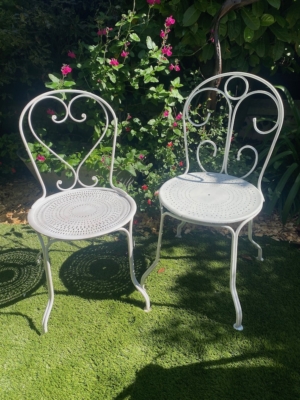 Deux chaises en fer forgé 1950 en blanc sont placées sur une pelouse verte, entourées de fleurs roses et de plantes luxuriantes. Brocante 93
