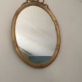 Le Miroir à entrelacs est un miroir ovale au cadre doré orné de motifs délicats, accroché à un mur blanc et reflétant une partie d'une pièce bleue. Brocante 93