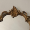Le Miroir Rococo 1960, doté d'un cadre doré orné de détails complexes sur le dessus, respire l'élégance vintage et s'accroche gracieusement à un mur uni de couleur claire. Brocante 93