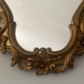 Le Miroir Rococo 1960 présente un cadre doré orné de détails floraux dans sa partie inférieure, reflétant un mur blanc uni. Brocante 93