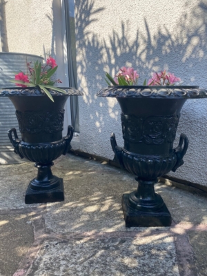 Une paire de vases Médicis XXème siècle, ornés de noir et remplis de fleurs roses, ornent un patio en pierre éclairé par le soleil à côté d'un mur de couleur claire, rayonnant d'une élégance intemporelle. Brocante 93