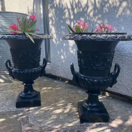 Une paire de vases Médicis XXème siècle, ornés de noir et remplis de fleurs roses, ornent un patio en pierre éclairé par le soleil à côté d'un mur de couleur claire, rayonnant d'une élégance intemporelle. Brocante 93