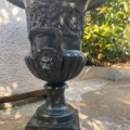 Une paire de vases Médicis XXème siècle, avec des motifs floraux et des visages, orne un patio en pierre éclairé par le soleil, à proximité de buissons verdoyants. Brocante 93