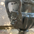 Gros plan d'un vase en métal de style Médicis de la Paire de vases Médicis XXème siècle, doté d'une anse ornée en forme de visage, présenté à l'extérieur contre un mur texturé. Brocante 93