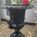 Paire de vases Médicis XXème siècle avec fleurs roses, posée sur une terrasse en pierre près d'un mur texturé et d'une fenêtre. Brocante 93