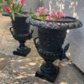 Une paire de vases Médicis XXème siècle en noir orné présente des fleurs roses sur un patio en pierre éclairé par le soleil, près d'un mur texturé. Brocante 93