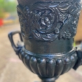 Gros plan sur une paire de vases Médicis XXème siècle, décor en métal noir avec gravures florales, posés en extérieur sur un fond flou et évoquant l'élégance intemporelle. Brocante 93