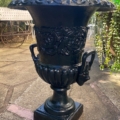 Paire de vases Médicis XXème siècle : urnes noires en métal orné de motifs floraux et de poignées, ajoutant une élégance classique à un patio rempli de plantes et de meubles de jardin. Brocante 93