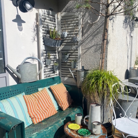 Une terrasse ensoleillée où trône la balancelle de jardin américaine des années 1950, agrémentée de coussins colorés, de plantes en pot et d'une petite table ronde dressée avec des tasses. Brocante 93