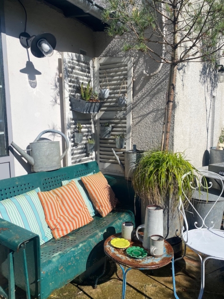 Une terrasse ensoleillée où trône la balancelle de jardin américaine des années 1950, agrémentée de coussins colorés, de plantes en pot et d'une petite table ronde dressée avec des tasses. Brocante 93