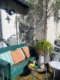 Une terrasse ensoleillée où trône la balancelle de jardin américaine des années 1950, agrémentée de coussins colorés, de plantes en pot et d'une petite table ronde dressée avec des tasses. Brocante 93