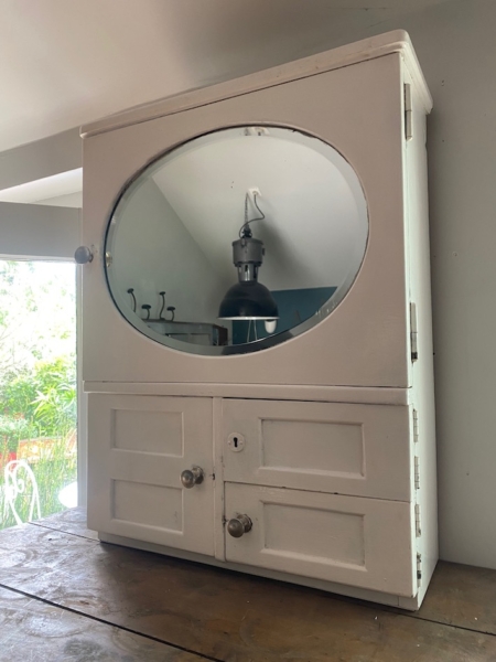 L'Armoire murale 1950, une armoire murale vintage blanche avec un miroir ovale, est posée sur une surface en bois près d'une fenêtre donnant sur une verdure luxuriante. Brocante 93