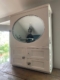 L'Armoire murale 1950, une armoire murale vintage blanche avec un miroir ovale, est posée sur une surface en bois près d'une fenêtre donnant sur une verdure luxuriante. Brocante 93