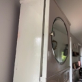 L'Armoire murale 1950, dotée d'un miroir rond sur la porte, est présentée en contre-plongée dans une pièce lumineuse et dégage un charme vintage subtil. Brocante 93