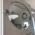 Le miroir rond sur une armoire blanche reflète le plafonnier, la fenêtre et le décor mural dans une pièce lumineuse dotée de l'Armoire murale 1950, qui lui confère un charme vintage. Brocante 93