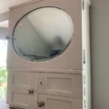 L'Armoire murale 1950 est une armoire vintage blanche avec un miroir ovale sur le dessus et deux portes à panneaux en dessous, idéale pour ajouter un charme rétro à n'importe quelle pièce. Brocante 93
