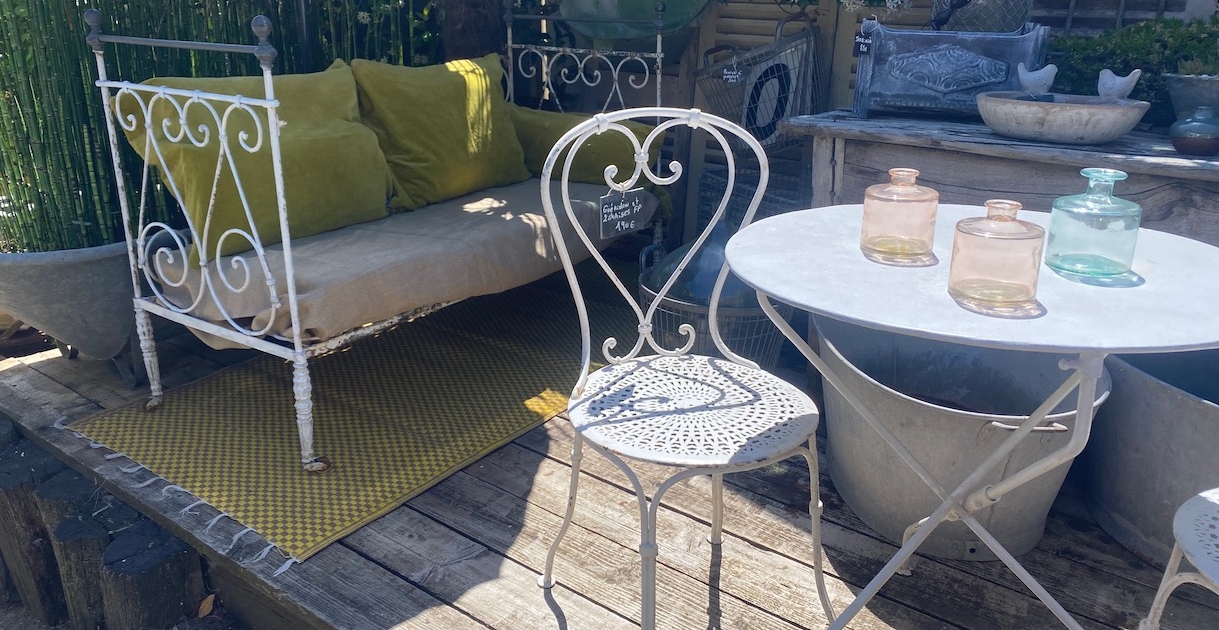 IMG_9690 Une terrasse au charme rétro avec une chaise en métal Carouche blanche, une table ronde, un lit de repos orné de coussins verts et des bocaux en verre posés sur une terrasse en bois. Brocante 93