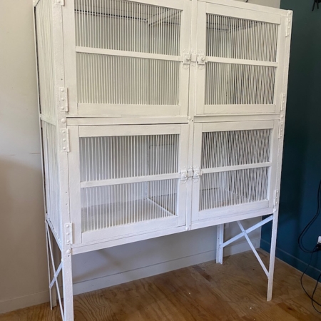 Le garde-manger vintage de 1950, une grande cage à oiseaux en métal blanc à quatre compartiments, se détache sur un mur bleu et blanc, posé sur un sol en bois. Brocante 93
