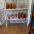 Vitrine vintage de garde-manger des années 1950, de couleur blanche, contenant des bouteilles remplies d'un liquide orange, des bocaux aux couvercles rouges et de la verrerie, le tout disposé sur un fond en bois. Brocante 93