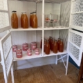 Le meuble « Garde Manger Vintage 1950 » est orné de bouteilles en verre contenant un liquide orange et de bocaux rouges et blancs, posés sur un plancher en bois, pour un charme rétro intemporel. Brocante 93