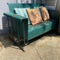 La balancelle de jardin américaine des années 1950, une balancelle en métal vert dotée de coussins rayés, trône sur une terrasse ensoleillée et patinée par les intempéries, évoquant le charme vintage américain. Brocante 93