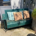 La « Balancelle de jardin américaine 1950 » est un banc en métal vert doté de trois coussins rayés, idéal pour se détendre avec élégance sur une terrasse ensoleillée. Brocante 93