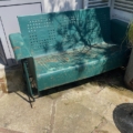 La balancelle de jardin américaine des années 1950, un siège en métal vert patiné, trône sur une terrasse ensoleillée et projette des ombres découpées sur le sol. Brocante 93