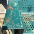 Gros plan sur une balancelle de jardin américaine des années 1950, patinée et de couleur vert rouille, dotée d'une assise et d'un dossier ajourés, baignée par la lumière du soleil à l'extérieur. Brocante 93