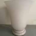 La lampe opaline Art déco 1930 est une lampe de table en verre opaline blanc de style Art déco, qui se distingue par son abat-jour large et évasé et son socle rond, évoquant l'élégance du design des années 1930 ; elle est présentée ici sur un support gris. Brocante 93
