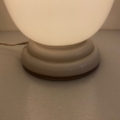 Une lampe Art déco de 1930 en opaline, allumée, dotée d'un socle blanc aux formes arrondies, repose sur une surface claire ; son cordon d'alimentation, bien visible, évoque l'élégance intemporelle du design des années 1930. Brocante 93