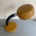 Une lampe berlinoise de 1970, dotée d'un abat-jour jaune et d'un col noir flexible au style rétro des années 1970, repose sur une surface gris clair devant un mur blanc. Brocante 93