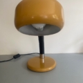 La « Lampe berlinoise 1970 », une lampe de bureau vintage jaune dotée d'un abat-jour rond, est posée sur une surface grise devant un mur blanc. Brocante 93