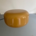 Une grosse meule ronde de fromage jaune repose sur une surface gris clair, devant un fond blanc uni, évoquant la lueur caractéristique de la « Lampe berlinoise 1970 ». Brocante 93