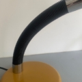 Lampe berlinoise de 1970, de couleur jaune, dotée d'un col flexible noir, présentée sur une surface grise avec un fond blanc. Brocante 93
