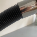 Gros plan sur un tuyau noir nervuré relié à un raccord métallique brillant, évoquant les éléments de design industriel de la « Lampe berlinoise 1970 », sur fond clair. Brocante 93