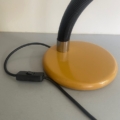 Lampe berlinoise de 1970 : un pied de lampe de bureau jaune, doté d'un bras flexible noir et d'un interrupteur sur le cordon, posé sur une surface grise — un design classique. Brocante 93
