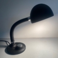 La « Lampe berlinoise 1970 » noire, dotée d'un col flexible et d'un socle rond, est allumée et éclaire une surface blanche. Brocante 93