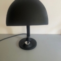 La « Lampe berlinoise 1970 », une lampe de table noire en forme de dôme dotée d'un pied rond et inspirée du style classique des années 1970, repose sur une surface gris clair devant un mur blanc. Brocante 93