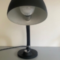 Une lampe berlinoise de 1970, au fini noir, dotée d'un pied rond et d'un abat-jour en forme de dôme, trône sur une surface grise devant un mur uni, incarnant tout le charme intemporel du design vintage des années 1970. Brocante 93