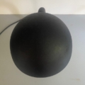 Une « Lampe berlinoise » noire de 1970, de forme bombée, munie d'un cordon, vue d'en haut sur une surface gris clair, à côté d'un mur blanc. Brocante 93