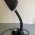 Lampe berlinoise de 1970, avec une finition noire, un bras flexible et un pied rond, posée sur une surface grise devant un mur blanc, dégageant un charme vintage subtil qui rappelle le design classique des années 1970. Brocante 93