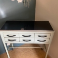 Le « Meuble de dentiste 1950 », une table en métal dotée de six tiroirs, d'un plateau noir et de pieds courbés, trône sur un parquet près du mur, apportant une touche de charme vintage à la pièce. Brocante 93