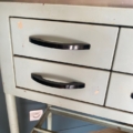 Une armoire vintage de dentiste datant des années 1950, dotée de deux tiroirs aux poignées noires et fabriquée en métal clair, est posée à côté d'une prise murale, sur un parquet. Brocante 93