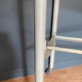 Gros plan sur un pied de table en métal dont la peinture est écaillée, rappelant le « Meuble de dentiste 1950 », placé près d'un mur bleu et d'un parquet. Brocante 93