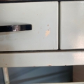 Gros plan sur un tiroir métallique vert pâle muni d'une poignée noire et présentant de petites écaillures de peinture, caractéristiques du « Meuble de dentiste 1950 ». Brocante 93