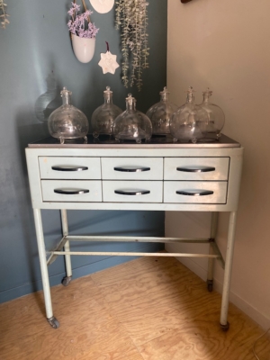 Un « Meuble de dentiste 1950 » — une armoire vintage en métal dotée de six tiroirs et surmontée de bocaux en verre — est posé contre un mur bleu et blanc. Brocante 93