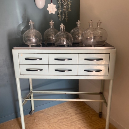 Un « Meuble de dentiste 1950 » — une armoire vintage en métal dotée de six tiroirs et surmontée de bocaux en verre — est posé contre un mur bleu et blanc. Brocante 93