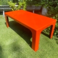 La « Table basse 1970 », une table basse rectangulaire de couleur orange vif, trône sur l'herbe verte d'un jardin entouré de plantes, avec un banc gris en arrière-plan. Brocante 93