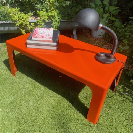 Une table basse orange vif datant des années 1970, ornée de livres et d'une lampe noire, posée sur l'herbe dans un décor de jardin, apporte une touche de charme vintage qui rappelle le design emblématique des années 1970. Brocante 93