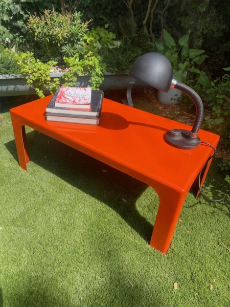 Une table basse orange vif datant des années 1970, ornée de livres et d'une lampe noire, posée sur l'herbe dans un décor de jardin, apporte une touche de charme vintage qui rappelle le design emblématique des années 1970. Brocante 93